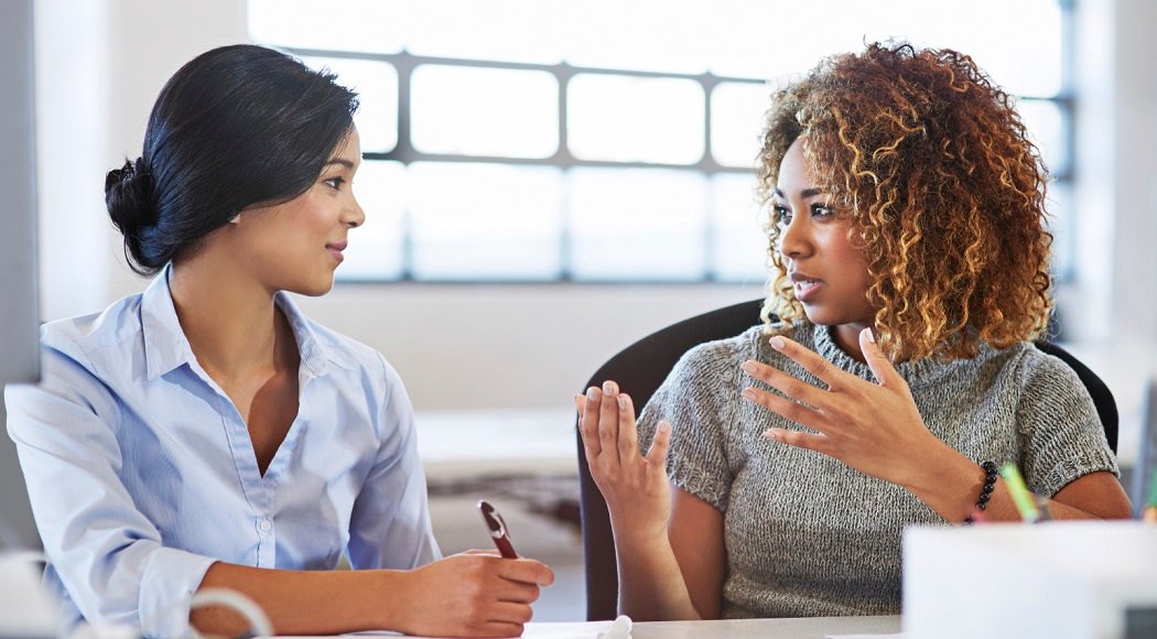 Two women engaged in a leadership coaching session at a table, with one listening attentively while the other gestures as she speaks.