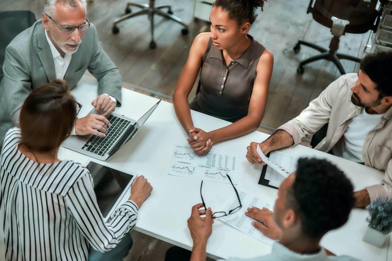 A group of people, led by a business coach, sitting around a table in an office.